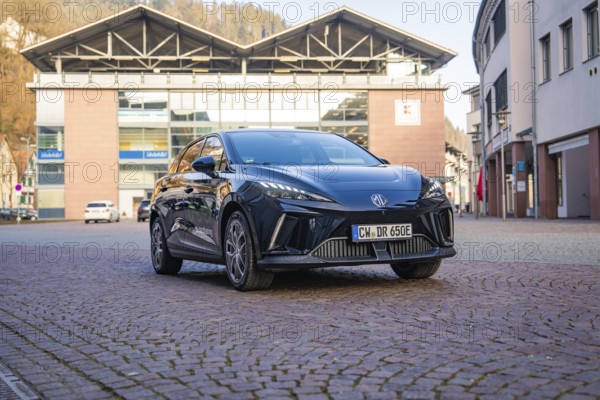 Black car parked in front of modern buildings on a paved area, illuminated by the sun, MG4 electric car, Deer e-Carsharing, Calw, Germany