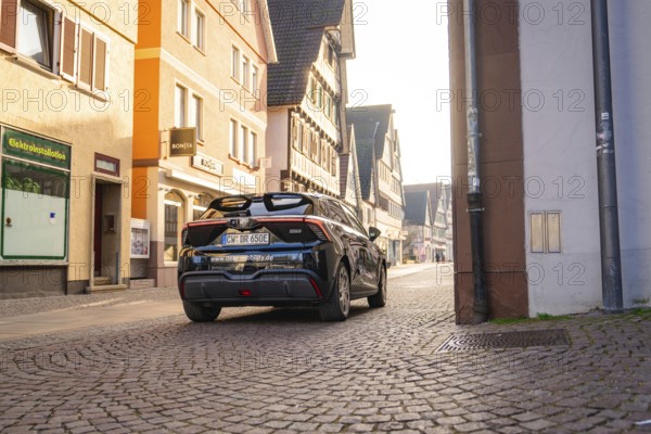 Black car drives through a sunny street lined with historic buildings, MG4 electric car, Deer e-Carsharing, Calw, Germany