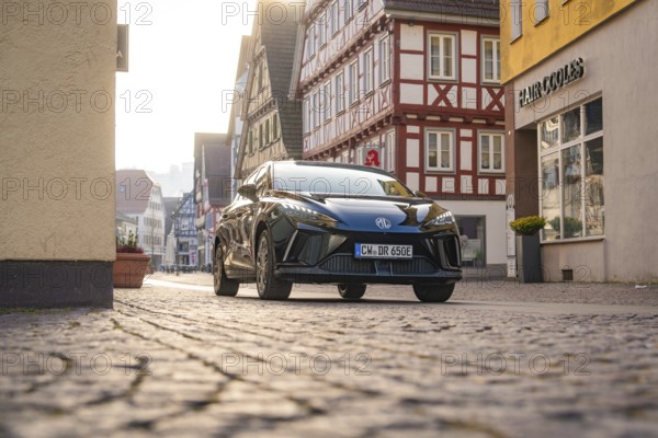 Black car drives through a paved street in a historic town with half-timbered houses, illuminated by sunlight, MG4 electric car, deer e-car sharing, Calw, Germany