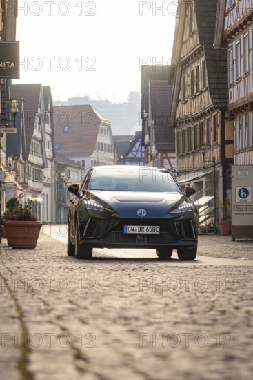 Black car on an empty, paved road surrounded by traditional building facades in sunshine, MG4 electric car, Deer e-Carsharing, Calw, Germany