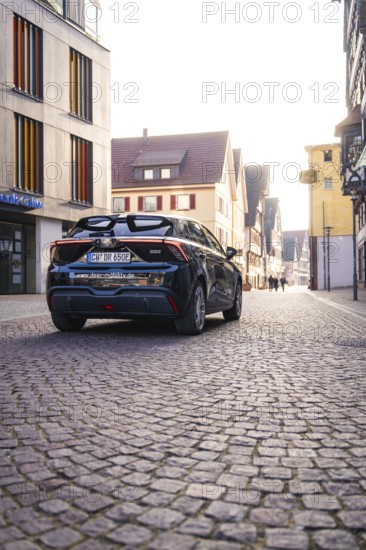 Black car drives through a paved city street between modern and historic buildings in sunlight, MG4 electric car, Deer e-Carsharing, Calw, Germany
