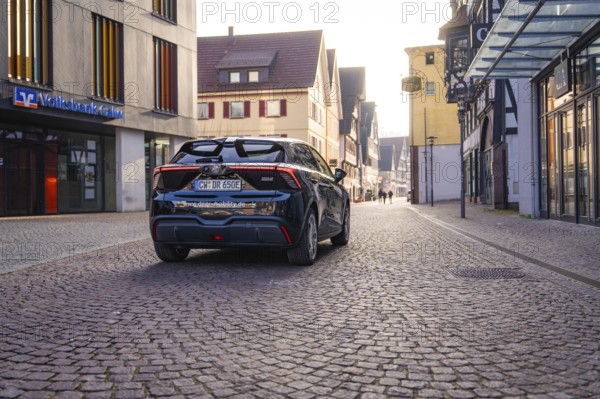 Black car on a paved street in the city with modern and historic architecture in the background at dusk, MG4 electric car, deer e-car sharing, Calw, Germany