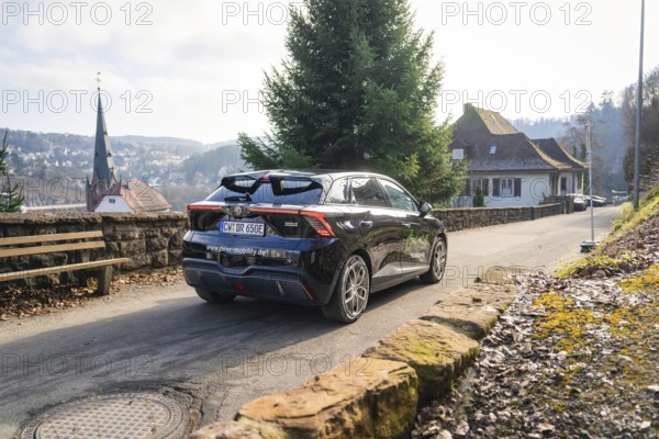 Car driving on a rural road with house and hills in the background, MG4 electric car, Deer e-Carsharing, Calw, Germany
