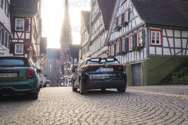 Timbered road with parked cars and church tower in the background in daylight, MG4 electric car, Deer e-Carsharing, Calw, Germany