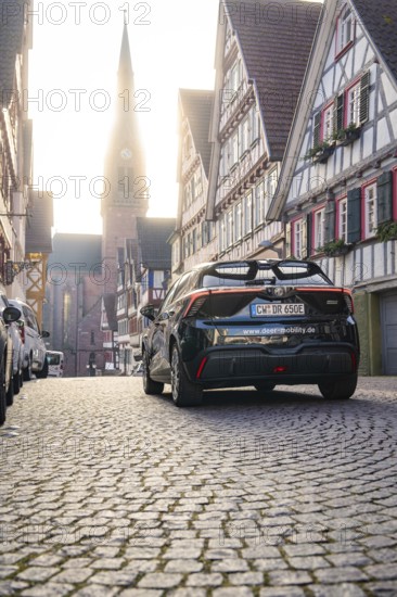 Car drives through a picturesque half-timbered road with church in the background at morning light, MG4 electric car, deer e-car sharing, Calw, Germany