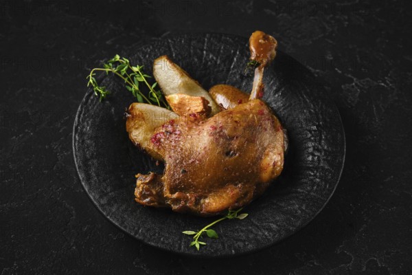 Duck confit on a black stone plate with pieces of roasted pear and herbs. The setting shows a dark background. The dish highlights the slow cooking method and texture of the meat