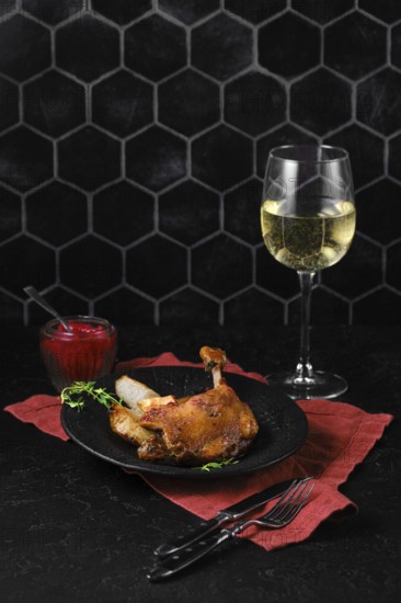 A plate with duck confit on a red cloth served with a glass of white wine and a lingonberry sauce