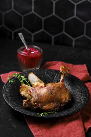 Roasted duck leg with crisp skin and tender meat on a stone plate. A small jar filled with a lingonberry sauce sits nearby. Green herbs are placed on the side of the duck on a dark table