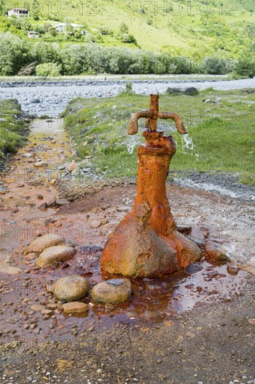 A rusty water spring over stones in front of a green landscape, mineral spring near Nadibani, White Aragvi River, Mtskheta-Mtianeti region, Georgian Military Highway, High Caucasus, Georgia