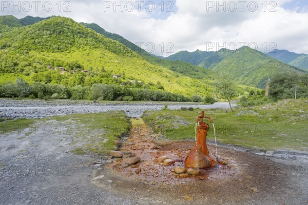 A rusty spring flows onto a gravel riverbed surrounded by green mountains, mineral spring near Nadibani, White Aragvi River, Mtskheta-Mtianeti region, Georgian Military Highway, High Caucasus, Georgia