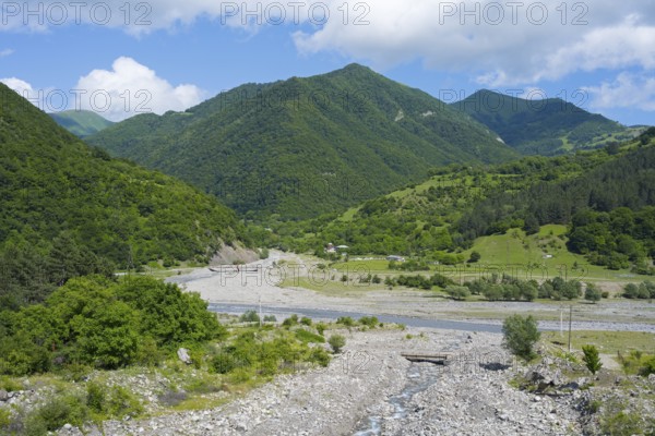 A riverbed leads through green mountains under a partly cloudy, blue sky, landscape near Chartali, White Aragvi River, Mtskheta-Mtianeti region, Georgian Military Highway, High Caucasus, Georgia