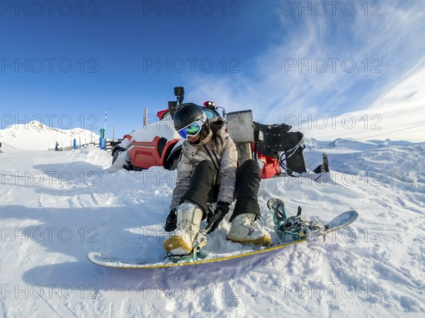 Young woman strapping her snowboard boots into the bindings on a snowy slope under a clear blue sky, preparing for a winter sport activity in a mountain resort