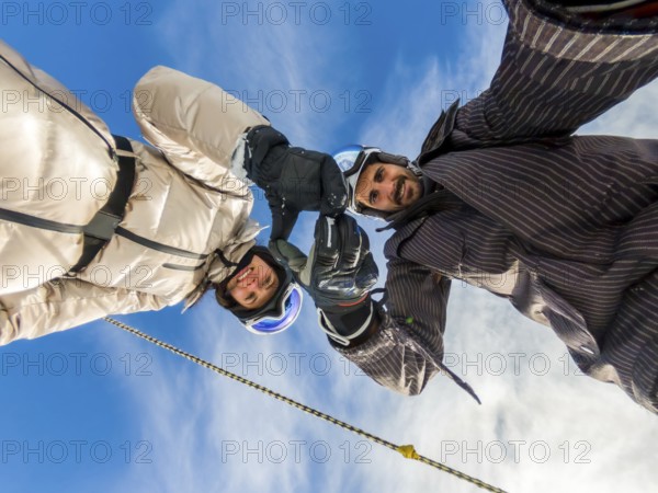 Happy couple wearing skiing gear and helmets, extending their hands upwards, forming an arch against a bright blue sky, enjoying winter sports adventure and freedom