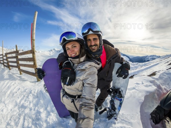 Couple smiling into camera while posing with snowboards on sunny snowy mountain slope, joyful selfie moment during a winter vacation at a ski resort, embracing and enjoying outdoor adventure
