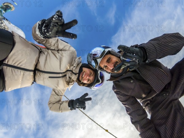Young couple smiling. Wearing ski helmets and goggles. Making peace signs and thumbs up gestures while taking a low angle selfie outdoors against a blue sky. Enjoying snowboard winter sports activity