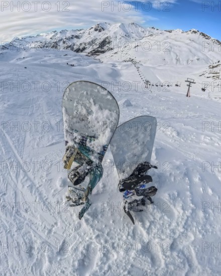 Snowboards rest in deep snow on a high mountain summit, offering a panoramic view of an expansive ski resort with distant peaks, snowy slopes, and a chairlift system under a bright winter sky