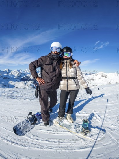 Snowboarders on a sunny mountain peak, smiling couple in helmets and goggles enjoying winter sports, snow covered panorama, clear blue sky and carefree adventure