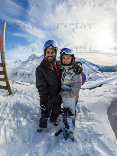 Joyful couple embracing on a snowy mountain peak, wearing helmets and goggles, holding a snowboard while enjoying their winter sports vacation under a blue sky