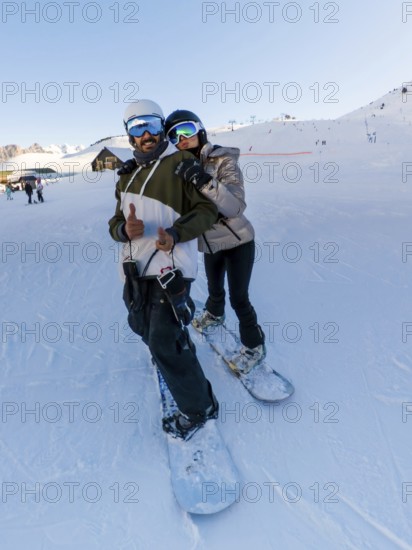 Couple wearing warm winter apparel and protective headgear smiling and posing for a selfie while standing with their snowboards on a professional ski slope under a clear sky