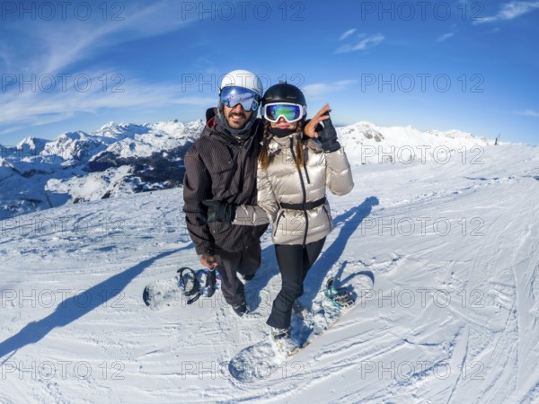 Couple posing with snowboards on an alpine peak, enjoying winter sports and a snowy mountain landscape under a bright blue sky, feeling happiness and adventure