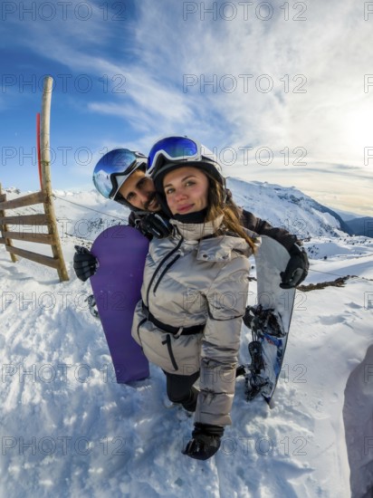 Young couple in helmets and goggles smile at camera, embracing with snowboards on a bright sunny winter day amid snowy mountain peaks, sharing adventure and joy