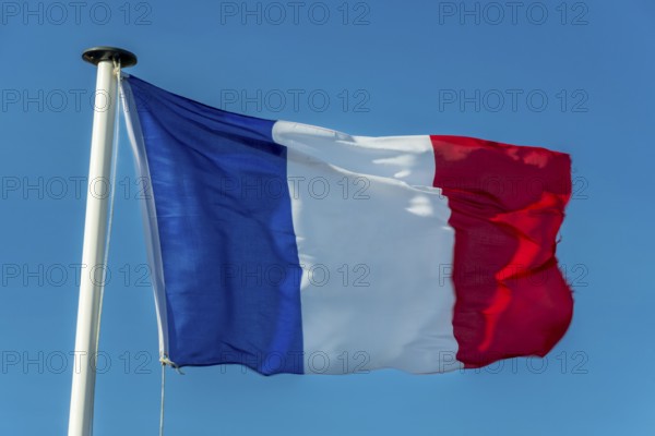 A French flag flutters in the wind on a white flagpole. The background is a clear, azure sky
