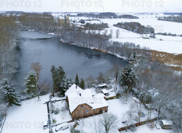 Winter landscape with a frozen lake and a snow-covered house on the shore, surrounded by forest, aerial view, Oelsburg, Ilsede municipality, Peine district, Lower Saxony, Germany