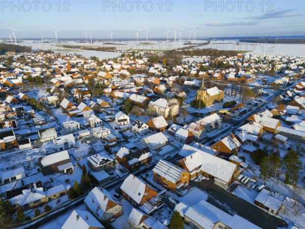 Snow-covered village with a church and a series of wind turbines on the horizon on a clear winter day, aerial view, Bernwardskirche, Groß Lafferde, municipality of Ilsede, district of Peine, Lower Saxony, Germany