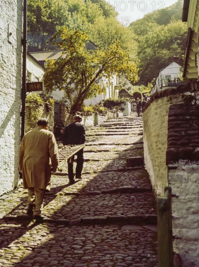 Men carrying wooden sled or sledge used for transport on steep hill of cobbled main village street, Clovelly, north Devon, England, UK 1960s