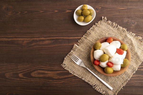 White Mozzarella cheese, with tomatoes and olives in wooden bowl on brown wooden background and linen textile, top view, flat lay, copy space