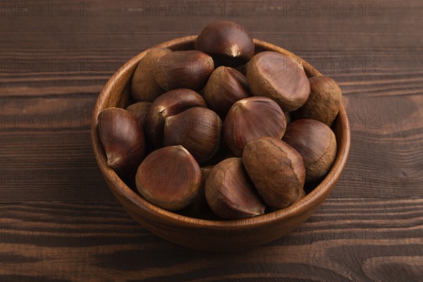 Wooden bowl with raw edible ?hestnuts on brown wooden background, side view, close up, minimalism