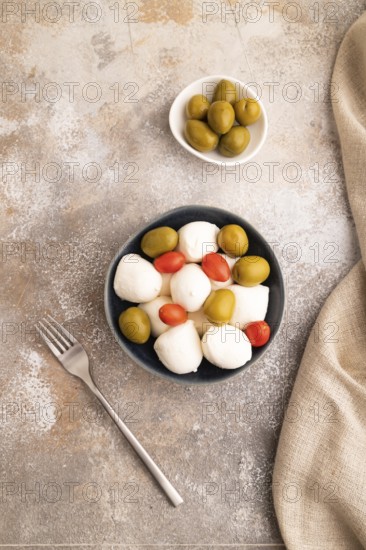 White Mozzarella cheese, with tomatoes and olives in blue bowl on brown concrete background and linen textile, top view, flat lay, close up