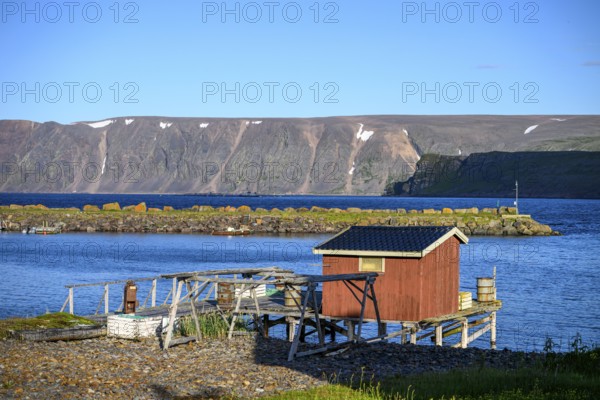 Small wooden cabin on a calm blue sea with mountain backdrop, red house on the shore with a view of the sea and rocks in the background, Nordfjord, Båtsfjord, Finnmark, Norway