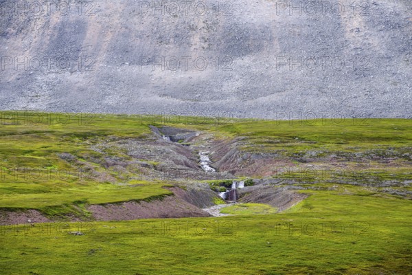 On the slope of a massive granite rock with rugged boulders at Austertanaveien 890 in the foreground, there is a torrent that cuts through green, flat fur beds, green low fell vegetation wide, quiet landscape, Tana, Finnmark, Norway