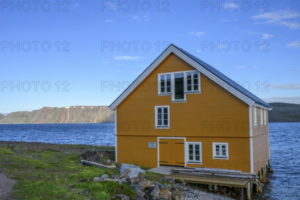 Yellow wooden house on the seashore under blue sky in natural landscape, yellow house overlooking the blue sea under a clear sky, Båtsfjord, Finnmark, Norway
