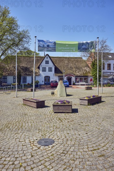 Advertising banners Welcome to the health resort of Leck!, ZumGlückgibtsleck, royally privileged pharmacy, Frisian house, general architecture, flagpoles, flower pots, boulder, trees with spring leaves, square of paving stones, pale blue sky, cirrostratus clouds, main street, market, leak, district of North Frisia, Schleswig-Holstein, Germany