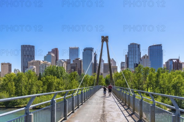 Two tourists are jogging across a pedestrian bridge in calgary, alberta with the downtown skyline visible in the background on a sunny summer day