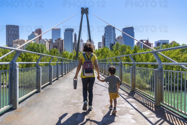 Mother and son holding hands, enjoying a sunny day walking on a bridge with the calgary city skyline in the background, showcasing urban life and family connection