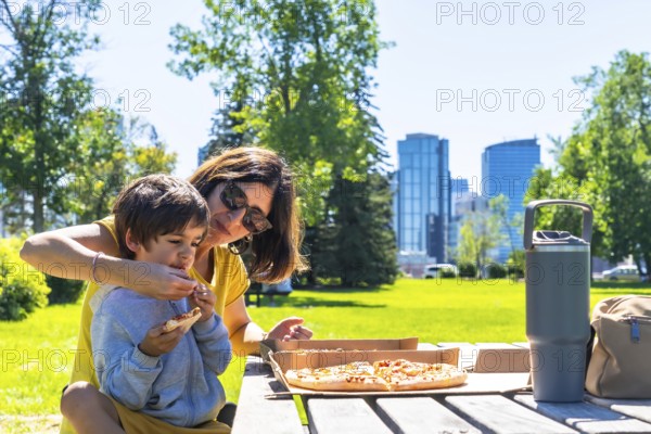 Mother helps her son eat a slice of pizza at a picnic table in a calgary park, enjoying quality time together on a beautiful sunny day with the city skyline in the background