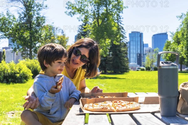 Mother and son are sharing a pizza picnic lunch together on a sunny day at a park in calgary, alberta, canada, with the city skyline in the background