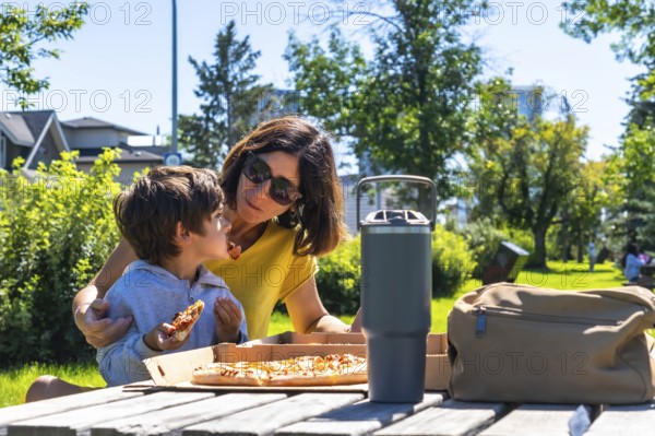 Mother and her son are enjoying a pizza picnic together on a sunny day in a calgary park, creating a heartwarming moment of family bonding