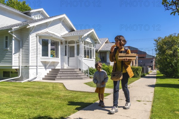 Mother and son are walking down a sidewalk in a residential neighborhood in calgary, alberta, canada, enjoying a pizza on a sunny day, with a white house in the background