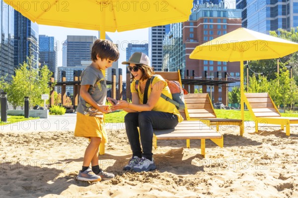 Mother and her son are spending quality time together at an urban beach in calgary, sheltered by yellow parasols, with the city skyline in the background