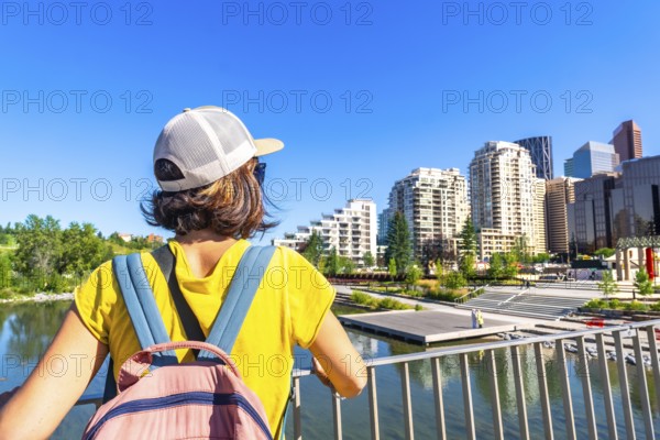 Tourist with a backpack is enjoying the cityscape of calgary, alberta, from a bridge, taking in the modern architecture and the bow river on a sunny day