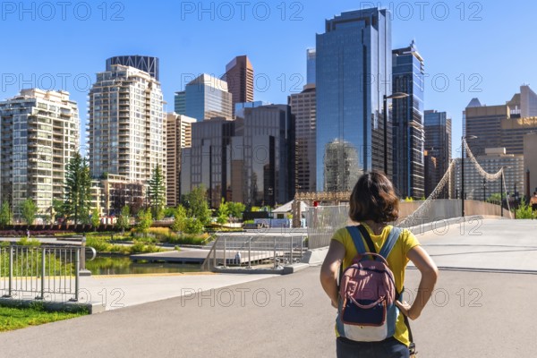 Young female tourist with a backpack is enjoying the cityscape of calgary, alberta, canada, standing on a bridge in a park with modern skyscrapers in the background on a bright summer day