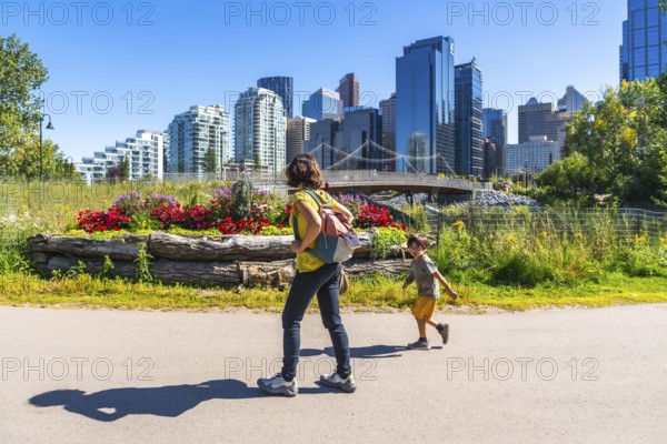 Mother with backpack walking with her running son in calgary's east village riverwalk, enjoying a sunny summer day with the city skyline in the background