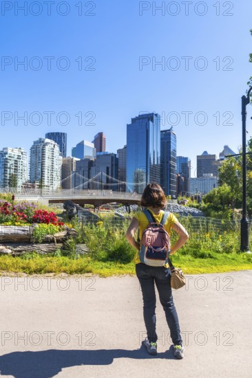 Tourist with a backpack stands in a colorful park, gazing at the modern cityscape of calgary, alberta, under a clear blue sky, enjoying the urban beauty and vibrant atmosphere