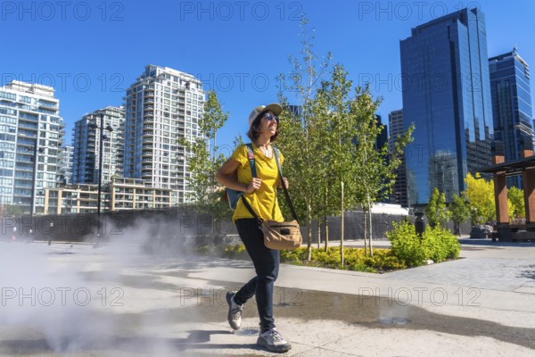 Female tourist walking through a refreshing misting station in calgary's east village, enjoying a sunny summer day while surrounded by urban skyscrapers and vibrant greenery