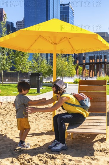 Mother and son enjoying a sunny day, playing together in a sandy urban park under a vibrant yellow beach umbrella, with calgary's skyline in the background