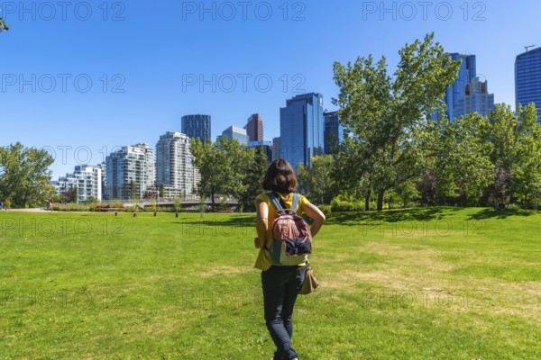 Female tourist with backpack walking on green grass in a park, enjoying the view of modern buildings of calgary's downtown under a clear blue sky on a sunny summer day